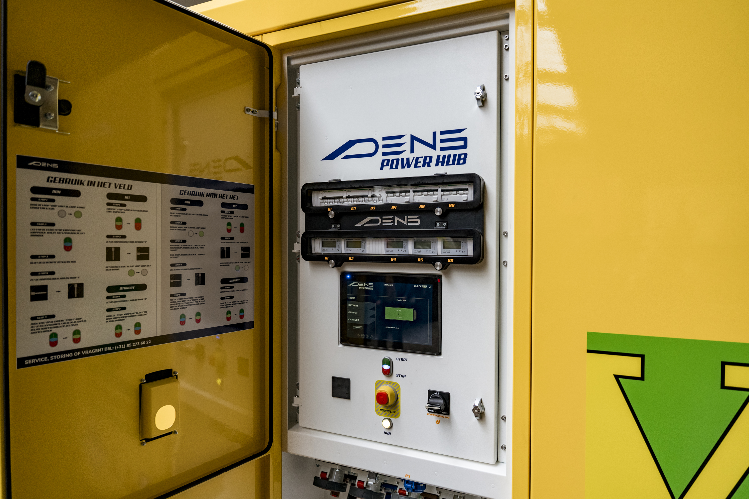 Het bedieningspaneel van de DENS Power Hub achter een geopende deur, met groepenkast, touchscreen en noodstop.